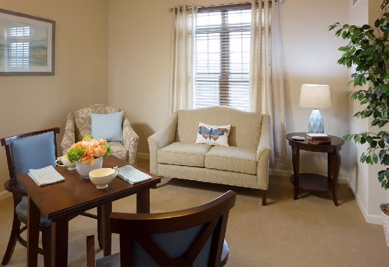 A cozy living room with a small dining table and chairs, an upholstered loveseat and armchair, side table with a lamp, and a potted plant.