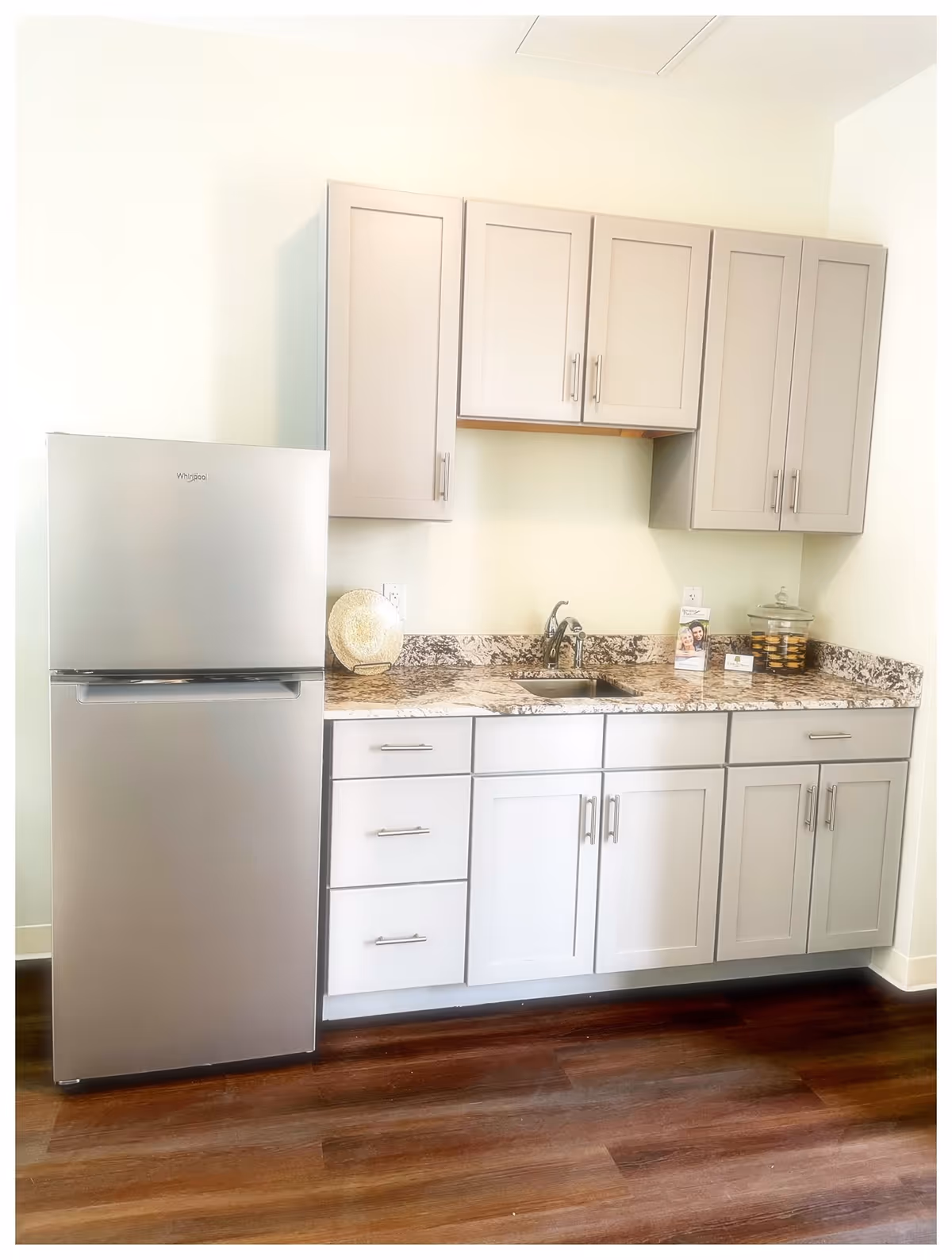 A modern kitchen area featuring a stainless steel Whirlpool refrigerator on the left, light gray cabinets above and below a granite countertop, a small sink with a faucet, and a few decorative items including a plate and a glass jar filled with cookies. The floor is a dark wood finish.