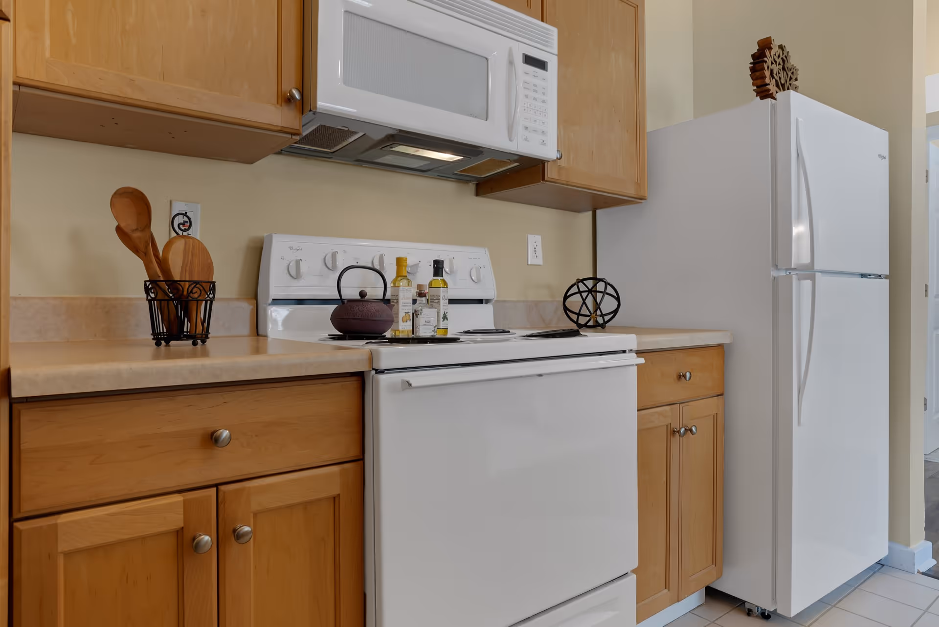 A kitchen area with light wood cabinets, a white stove with a black kettle and bottles on top, a white microwave above the stove, and a white refrigerator to the right. Wooden cooking utensils are in a black holder on the countertop to the left.