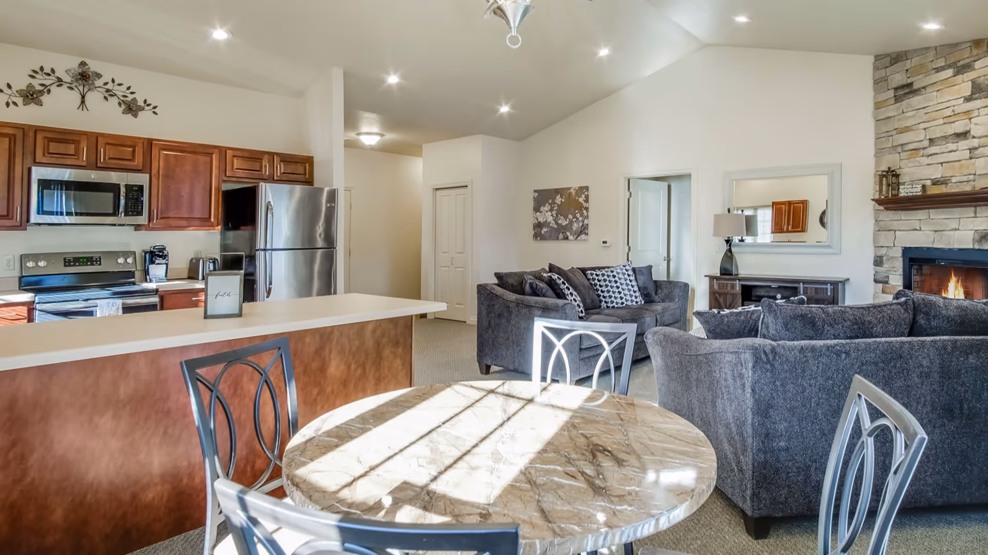 A bright and spacious open-concept living area featuring a kitchen with wooden cabinets, stainless steel appliances including a microwave and refrigerator, and a breakfast bar with a chair. In the foreground, there is a round marble table with metal chairs. The living room area has two gray sofas with patterned pillows, a stone fireplace with a fire burning, a side table with a lamp, and a wall mirror. The walls are white, and the ceiling is vaulted with recessed lighting.