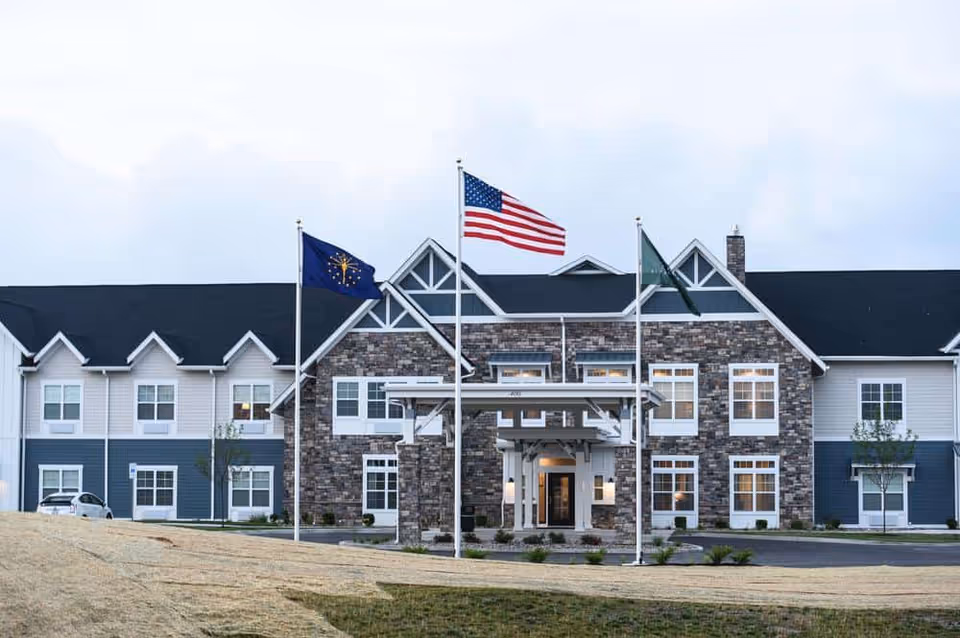 Front exterior of a two-story senior living building with a stone facade, covered entrance, and three flagpoles flying the American and state flags.