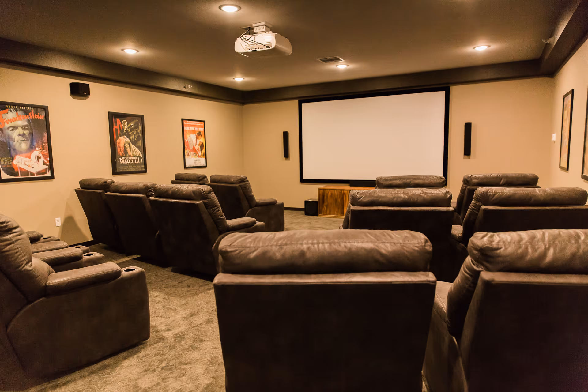 A cozy home theater room with multiple rows of dark brown recliner chairs facing a large white projection screen. The walls are beige and decorated with framed vintage horror movie posters including Frankenstein and Dracula. Ceiling lights and a projector are mounted overhead.