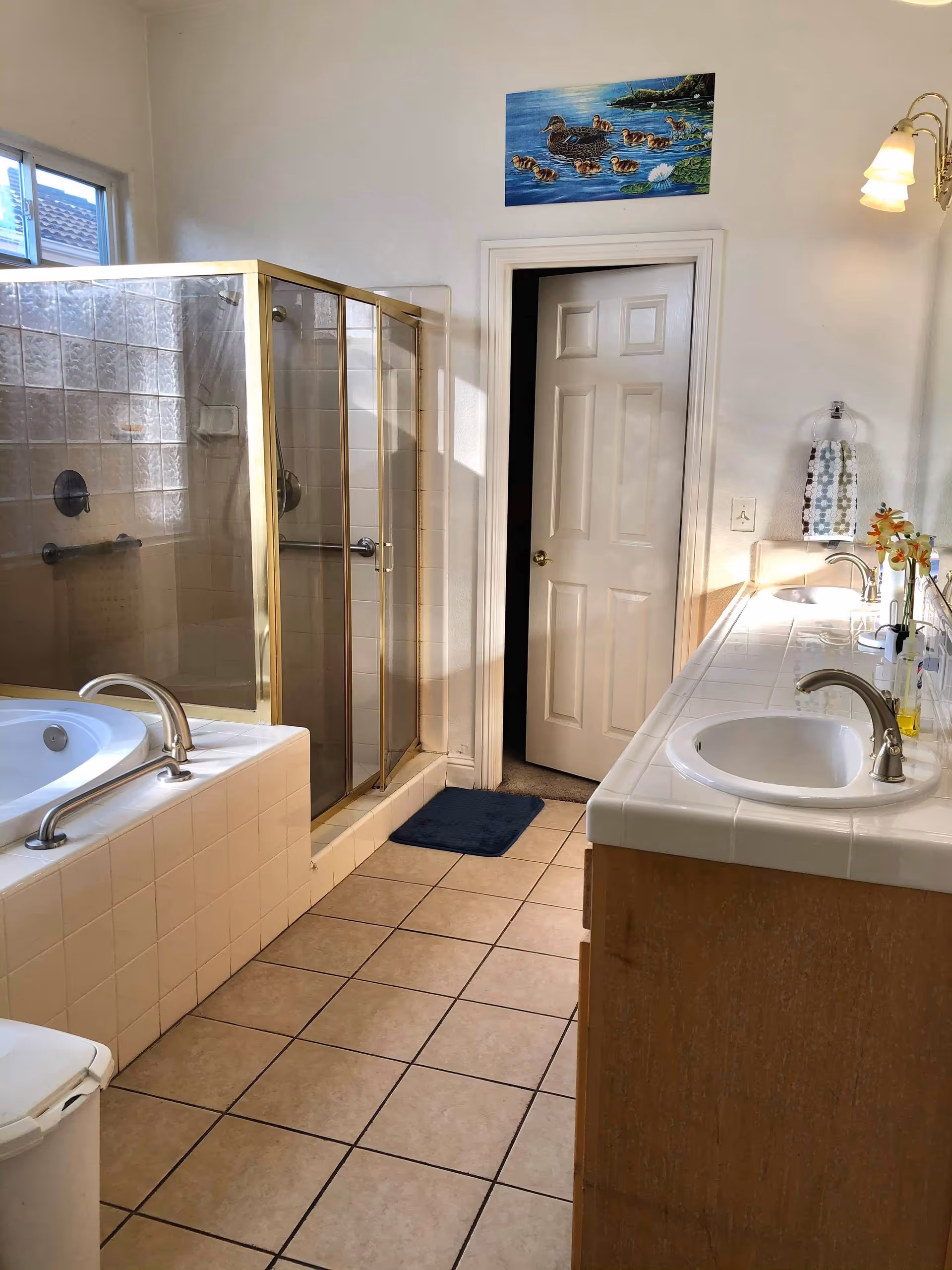 A bathroom with a tiled floor featuring a bathtub with a silver grab bar, a glass-enclosed shower with gold trim, a double sink vanity with white countertops and wooden cabinets, a towel hanging on the wall, and a painting of ducks above a white door.