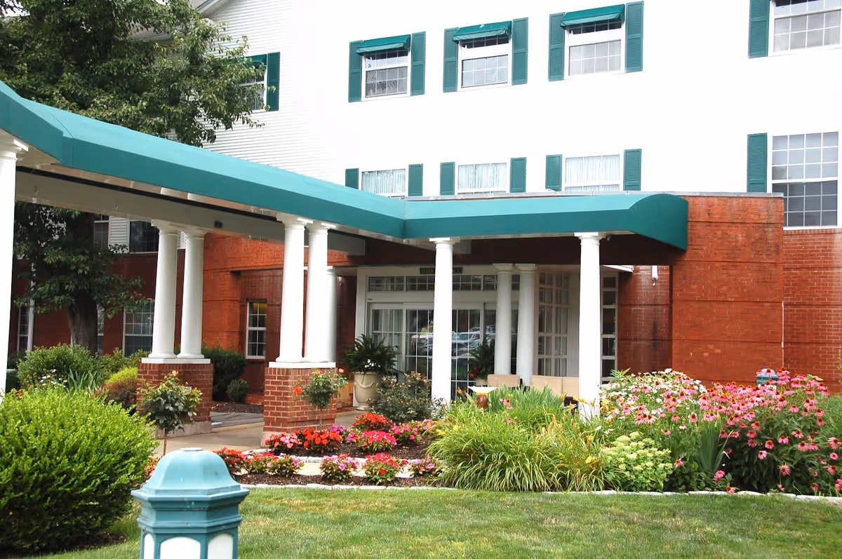 Entrance of a senior living facility with a covered drop-off area supported by white columns, surrounded by well-maintained flower beds and greenery. The building has white siding with green window shutters and red brick accents.