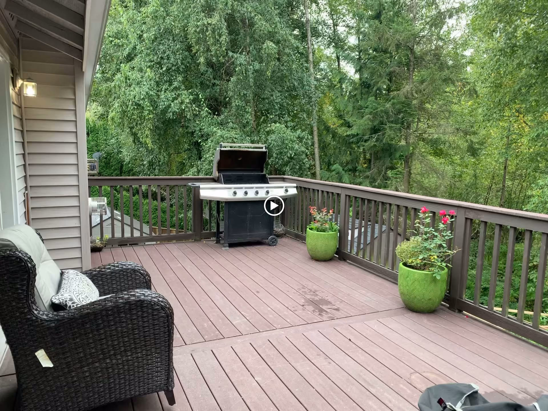 Outdoor wooden deck with a black wicker chair with cushions on the left, a gas grill with its lid open in the center, and two green planters with flowering plants along the railing. The deck overlooks a lush green forested area.