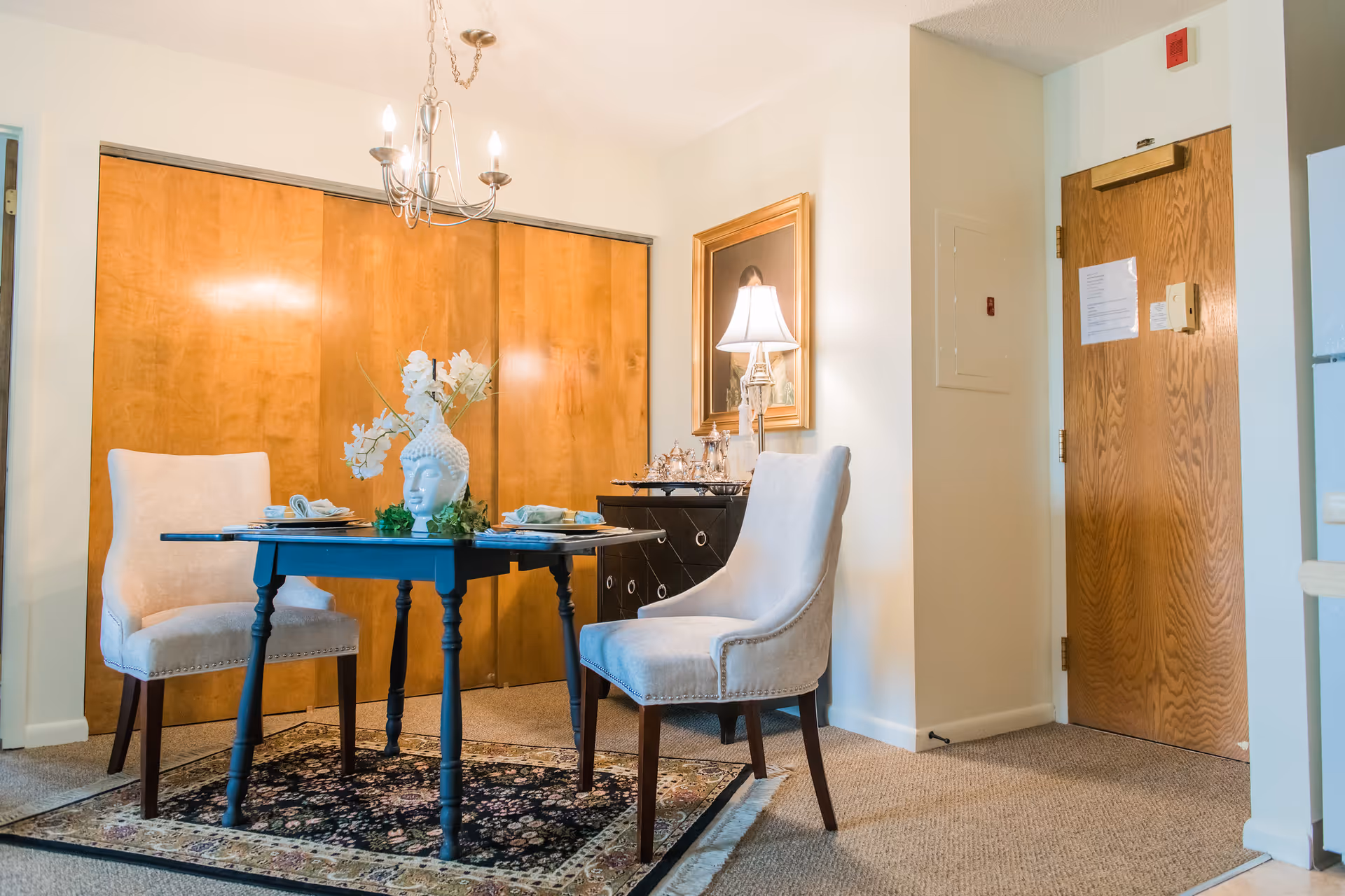 Small dining area with a black table set for two, upholstered chairs, a decorative centerpiece and lamp, and wooden closet doors in the background.