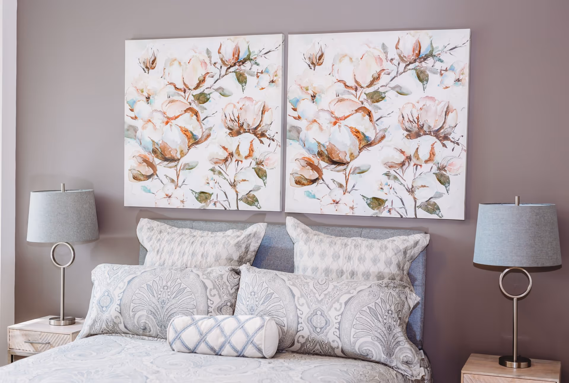 A neatly made bed with patterned pillows and a comforter in soft gray and white tones. Above the bed are two large floral paintings featuring white and brown flowers with green leaves. On each side of the bed is a wooden nightstand with a modern lamp that has a circular base and a gray lampshade. The wall behind the bed is painted in a muted mauve color.