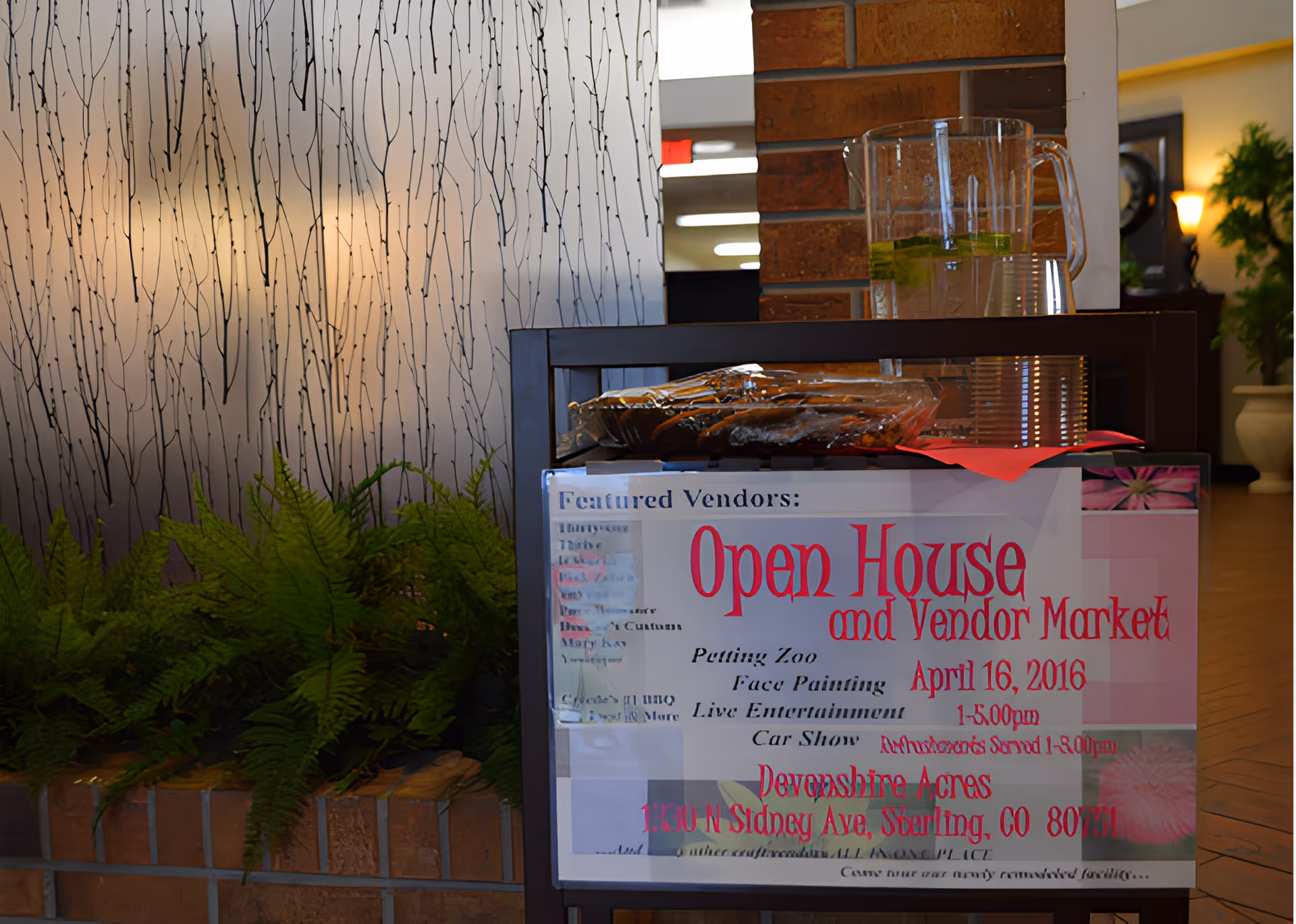 A sign advertising an Open House and Vendor Market event at Devonshire Acres, placed on a stand in an indoor setting with a brick wall and plants nearby. On top of the stand, there is a pitcher of water with lemon slices and a plate covered with plastic wrap. The background includes a decorative frosted glass panel and some indoor plants.