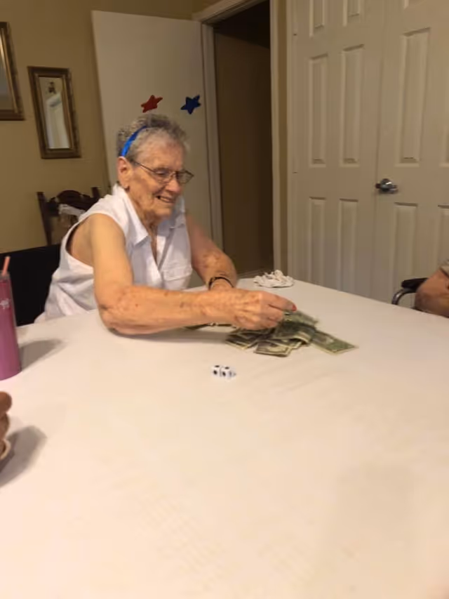 An elderly woman wearing a white sleeveless shirt and a headband with red and blue stars is sitting at a table counting money. There is a die on the table and another person partially visible on the right side. The room has beige walls, a door, and a mirror on the wall.