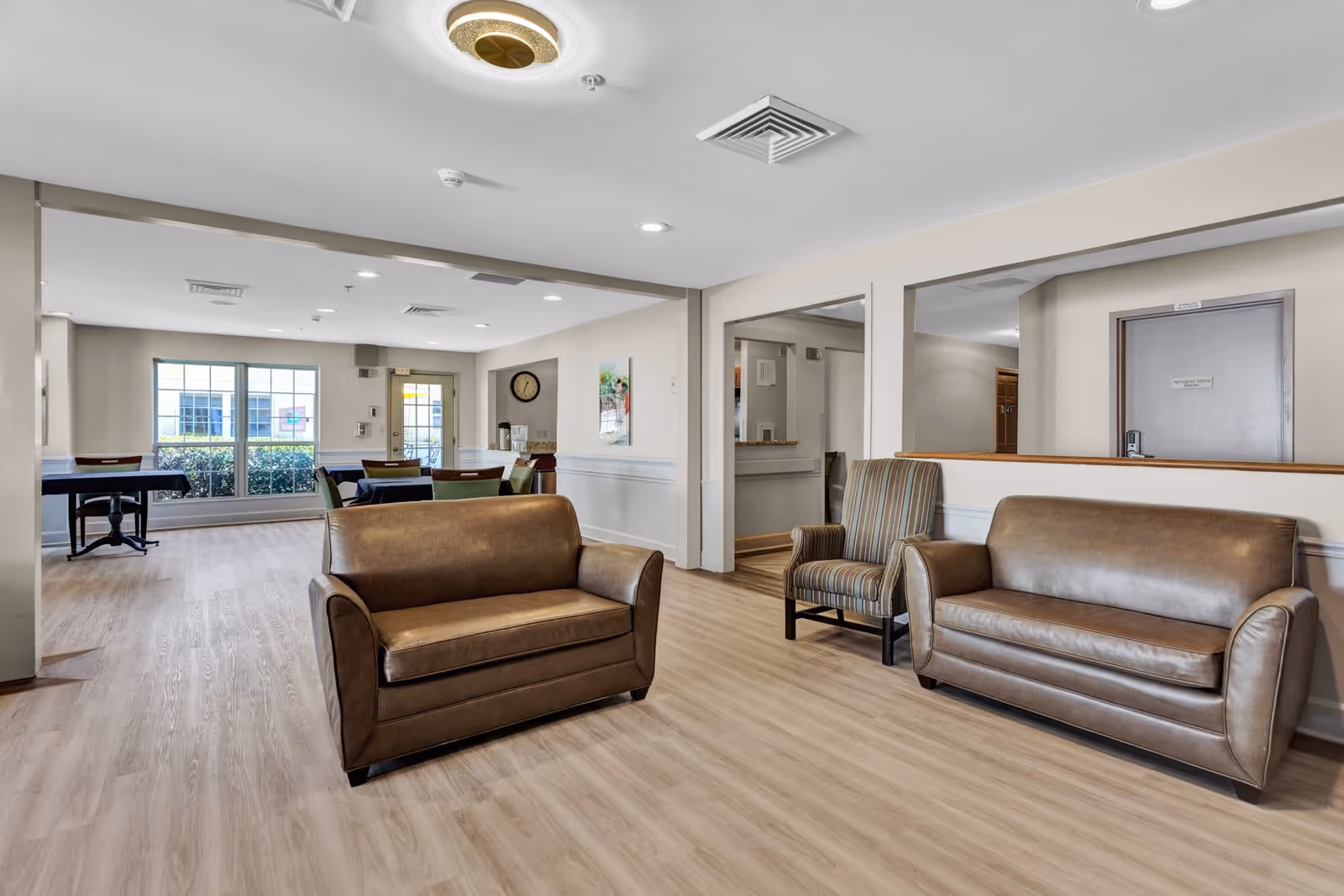 A bright and spacious common area in an assisted living facility featuring two brown leather sofas and a striped armchair on light wood flooring. In the background, there are tables with chairs near large windows letting in natural light, and a door leading outside. The walls are light-colored with white wainscoting, and there is a clock and artwork on the walls.