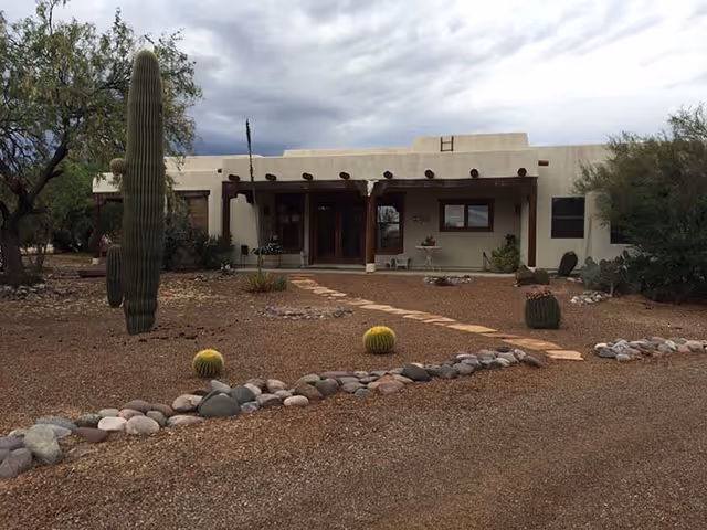 Single-story southwestern-style building with a covered front porch, desert landscaping, and cacti under a cloudy sky.