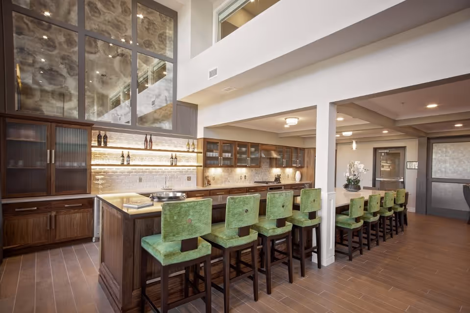 Spacious modern kitchen area with a long island counter featuring green cushioned bar stools. The kitchen has wooden cabinets with glass doors, a tiled backsplash with under-cabinet lighting, and a high ceiling with large windows reflecting the ceiling lights. There is a decorative plant on the counter and a door visible in the background.