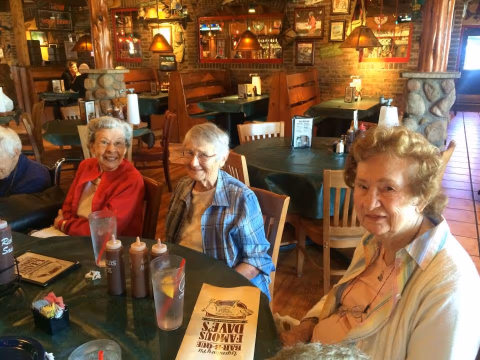 Three elderly women sitting around a table in a cozy restaurant with wooden furniture and stone pillars. The table has menus, glasses of water with lemon, and several bottles of sauces. The background shows more tables and warm lighting fixtures.