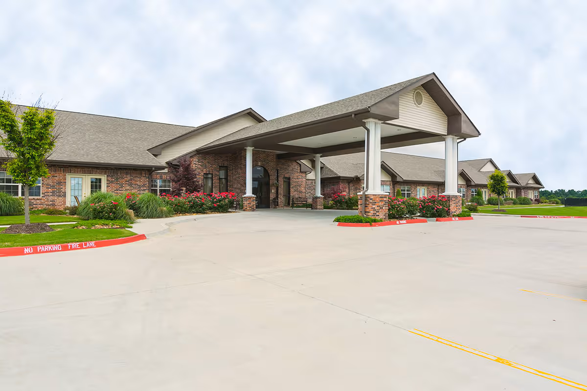 Exterior view of The Bungalows at Fayetteville showing a brick building with a covered entrance supported by white columns, surrounded by landscaped greenery and flowers, with a large concrete driveway in front.