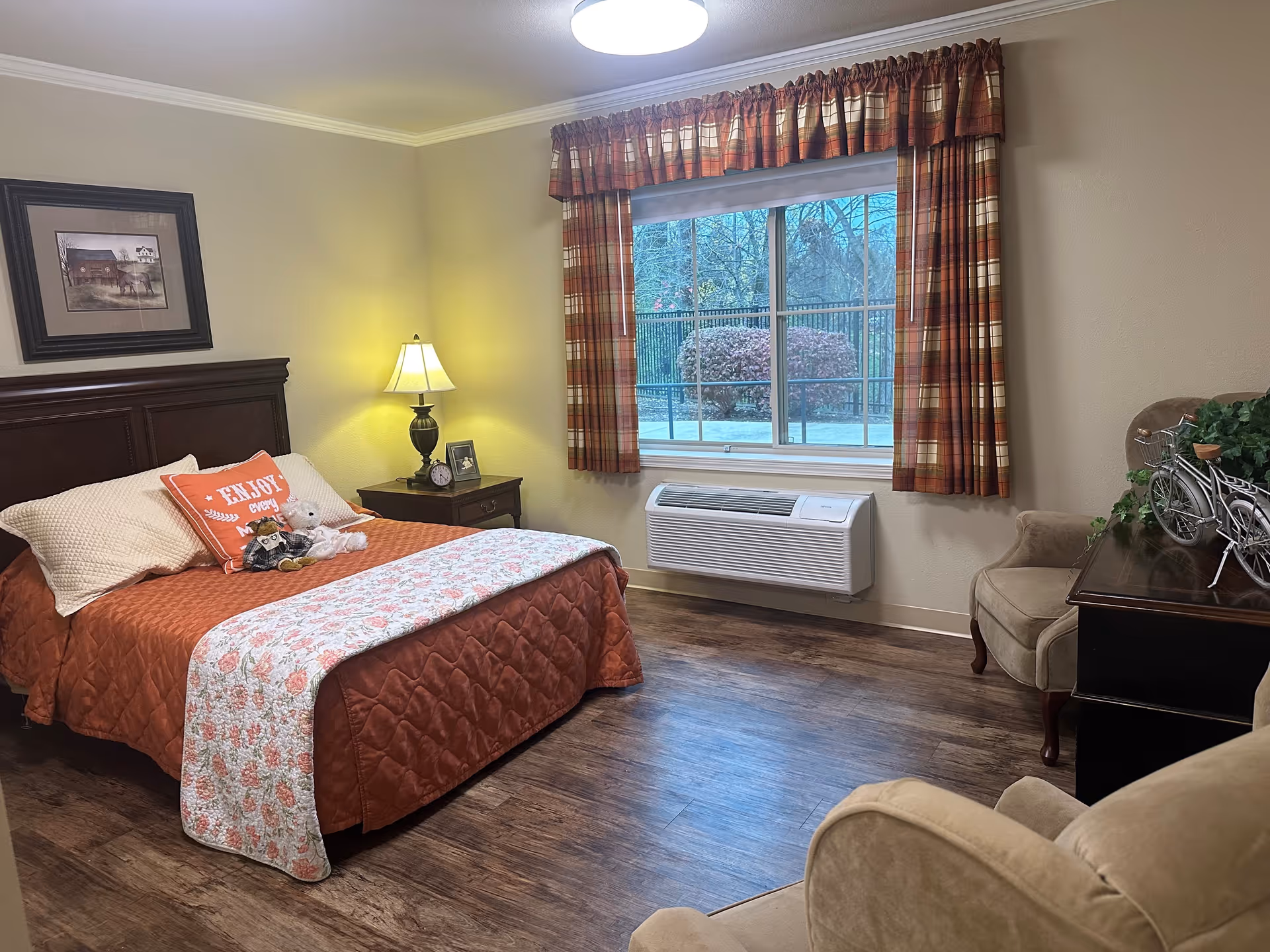 A cozy bedroom with a large bed covered in an orange quilt and a floral blanket at the foot. There are two pillows, one with the text 'ENJOY every moment'. A nightstand with a lamp, clock, and framed photo is beside the bed. A window with plaid curtains shows an outdoor view with bushes and a fence. The room has wooden flooring, a beige armchair, and a dark wooden dresser with a decorative bicycle and a plant on top.