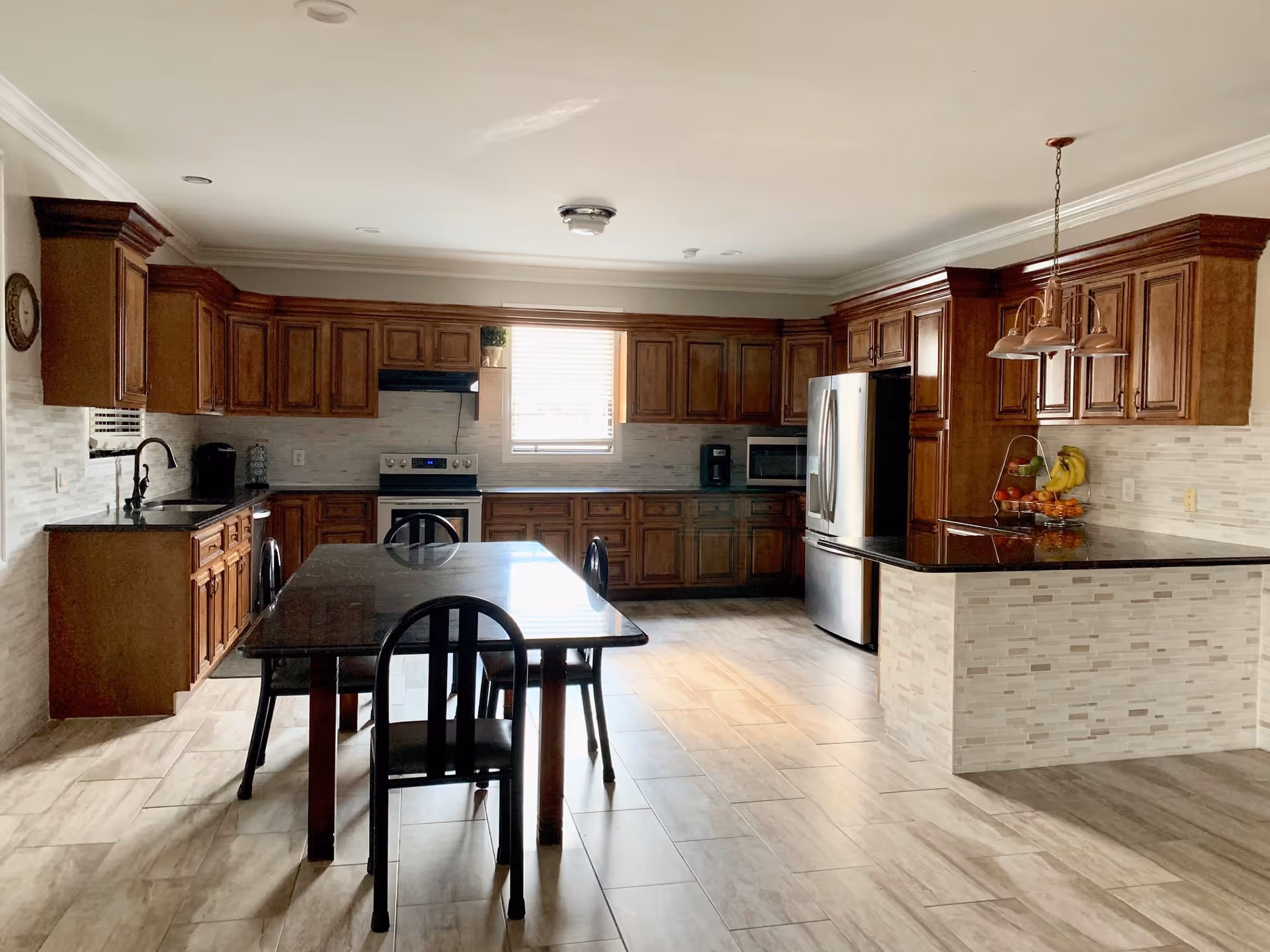 A spacious kitchen with wooden cabinets, a stainless steel refrigerator, stove, microwave, and coffee maker. There is a black dining table with four chairs in the center, and a kitchen island with a fruit basket on it. The floor is tiled, and a window with blinds lets in natural light.