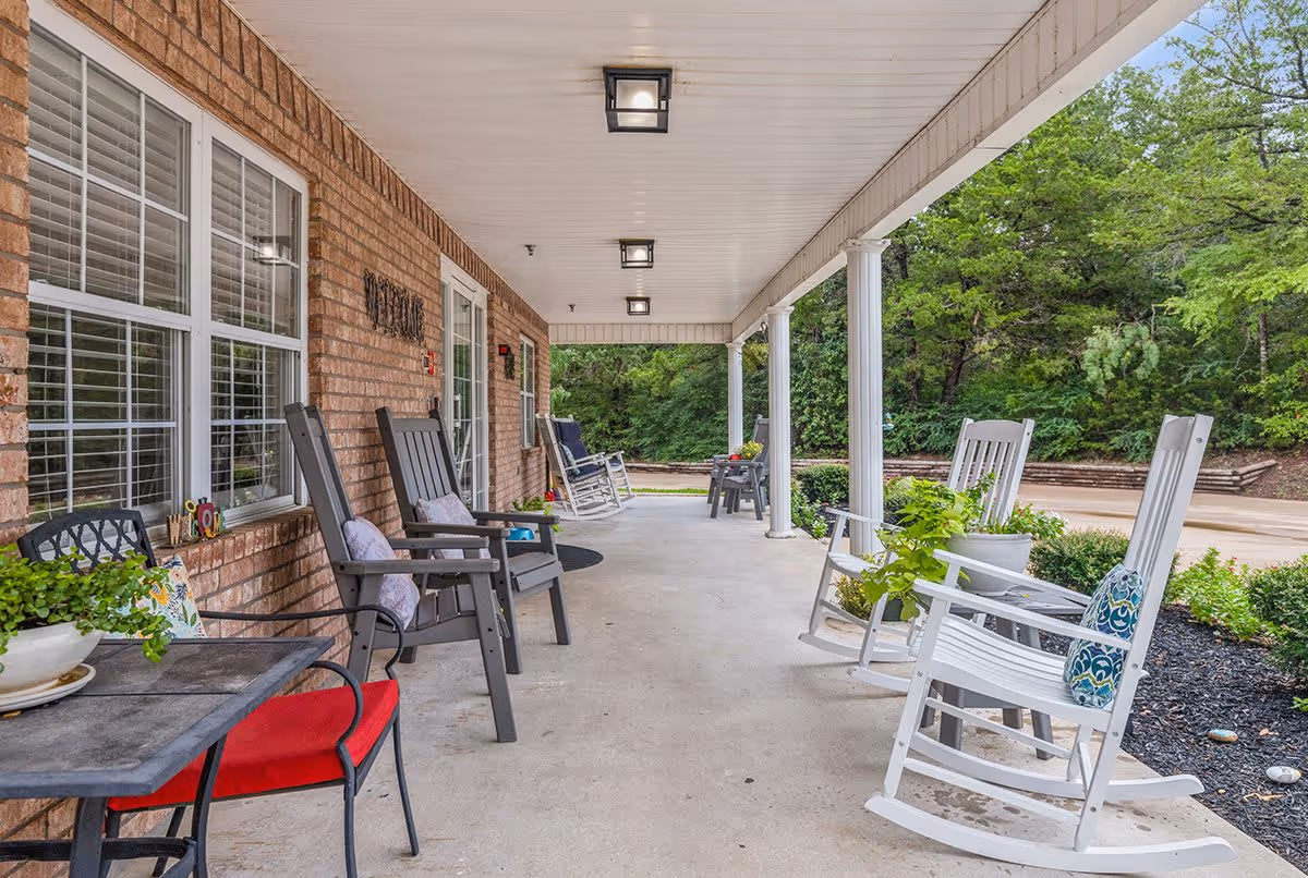 Covered outdoor porch area with several rocking chairs and a small table with a red cushioned chair. The porch has a brick wall on one side with windows and a door, and white columns supporting the roof. There are plants and greenery visible around the porch and in the background.