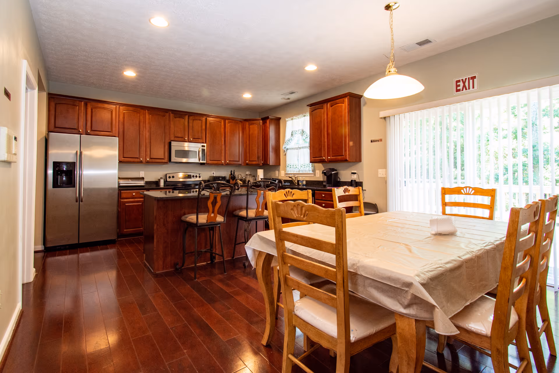 A spacious kitchen and dining area with wooden cabinets, stainless steel refrigerator, stove, microwave, and a kitchen island with bar stools. A dining table with six wooden chairs is covered with a beige tablecloth. Large vertical blinds cover a sliding glass door with an exit sign above it. The floor is dark hardwood and the ceiling has recessed lighting.