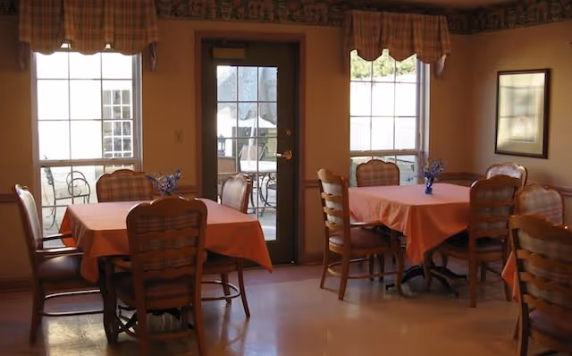 Small dining room with several tables covered in peach tablecloths, wooden chairs, and windows and a door leading to an outdoor patio.