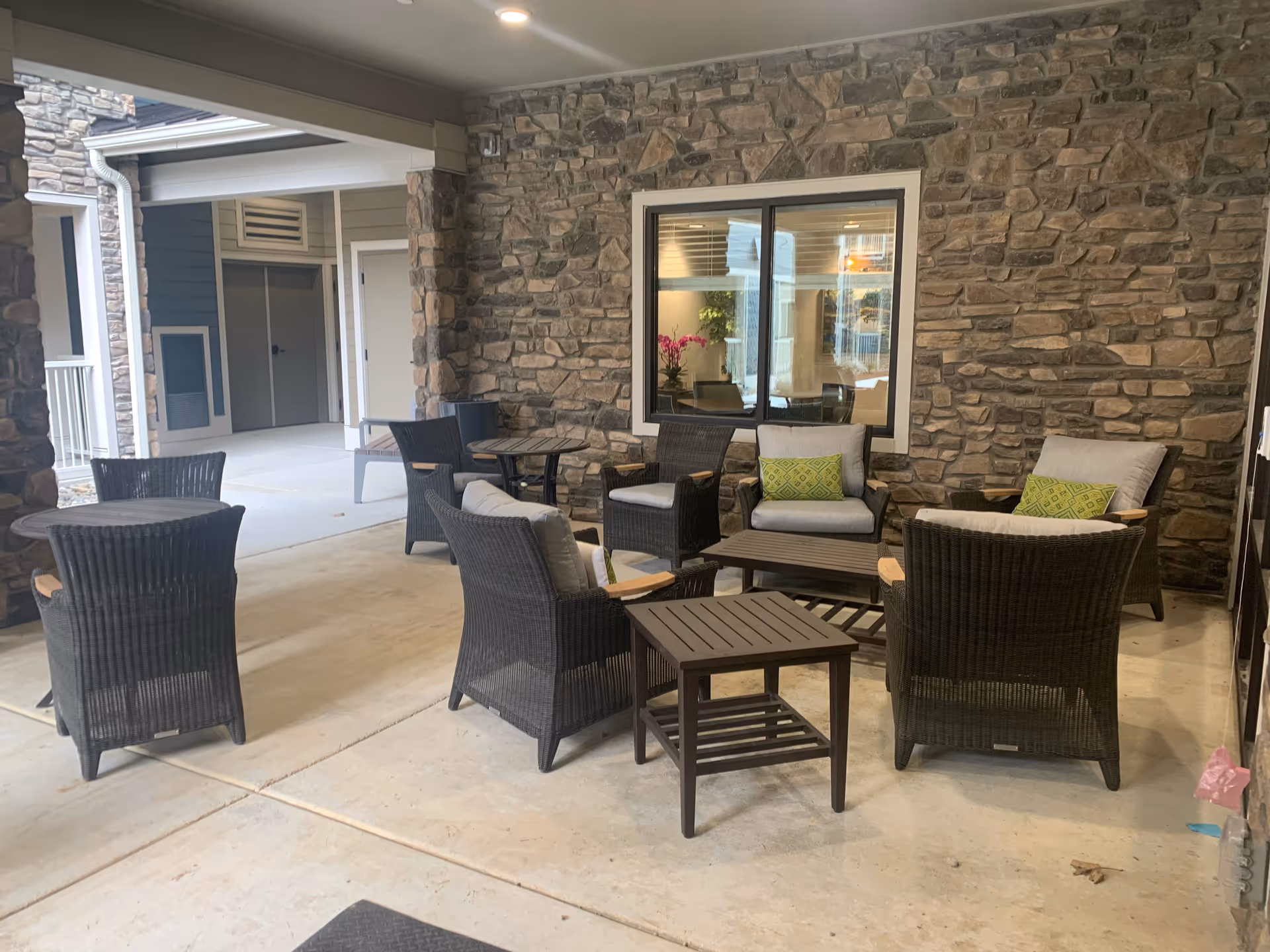 Covered outdoor seating area with several dark wicker chairs and tables arranged on a concrete floor. The background features a stone wall with a window showing an indoor room with plants and furniture.
