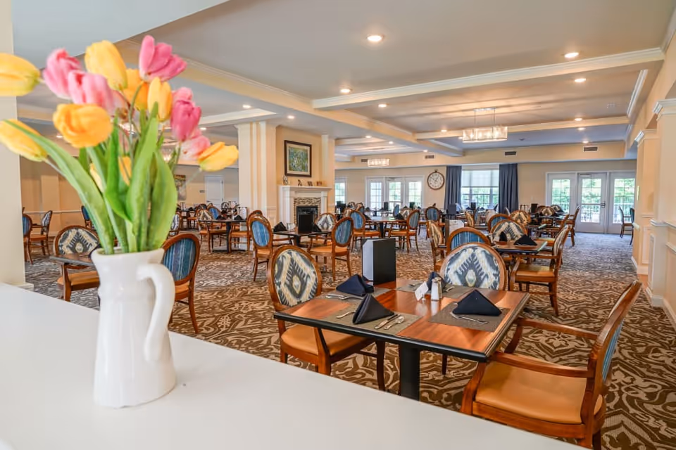 A bright dining room with multiple set tables and chairs and a vase of colorful tulips in the foreground.