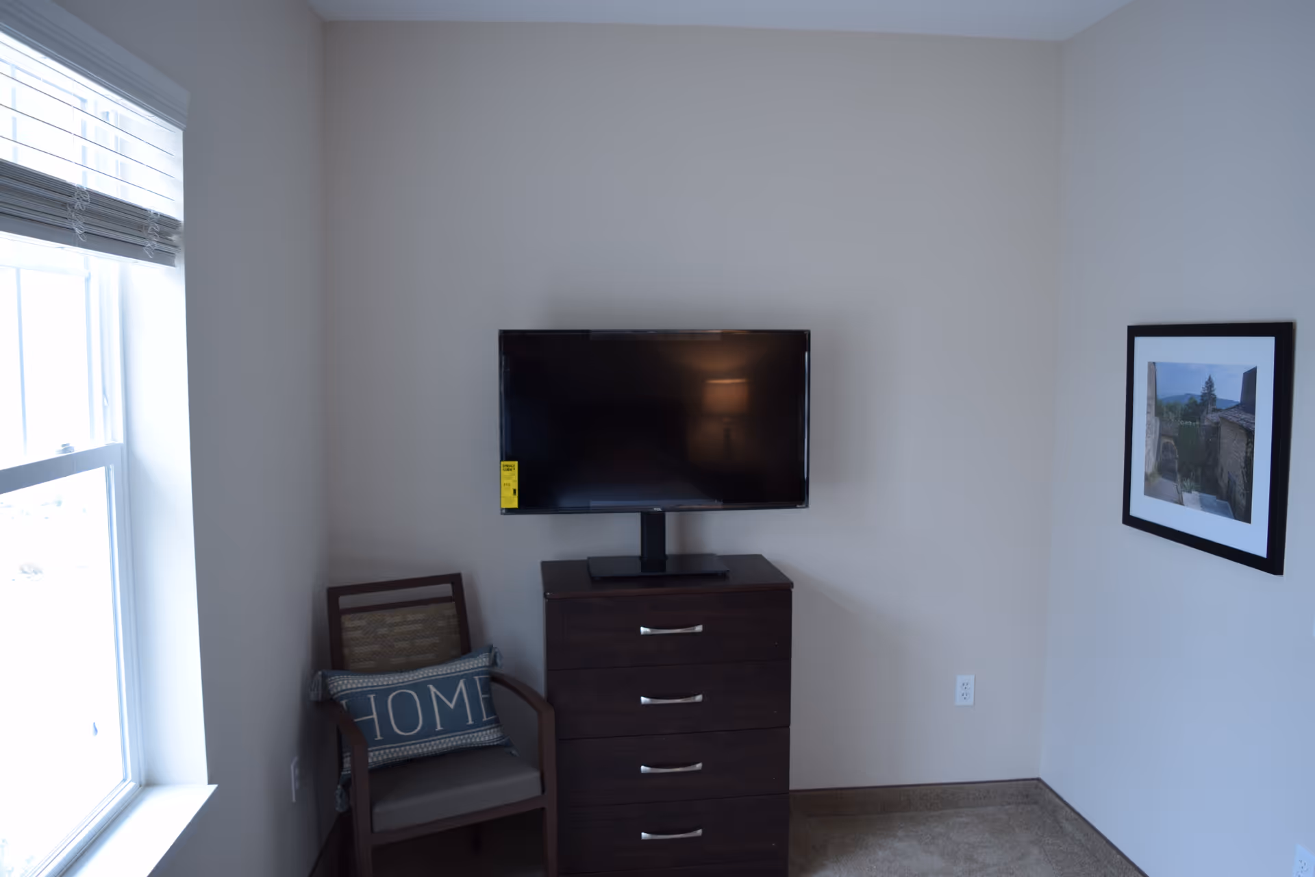 A corner of a room with a window on the left letting in natural light. Below the window is a wooden chair with a cushion that has the word 'HOME' on it. Next to the chair is a dark wooden dresser with four drawers, on top of which is a flat-screen TV mounted on a stand. On the wall to the right of the dresser is a framed picture of a scenic outdoor view.