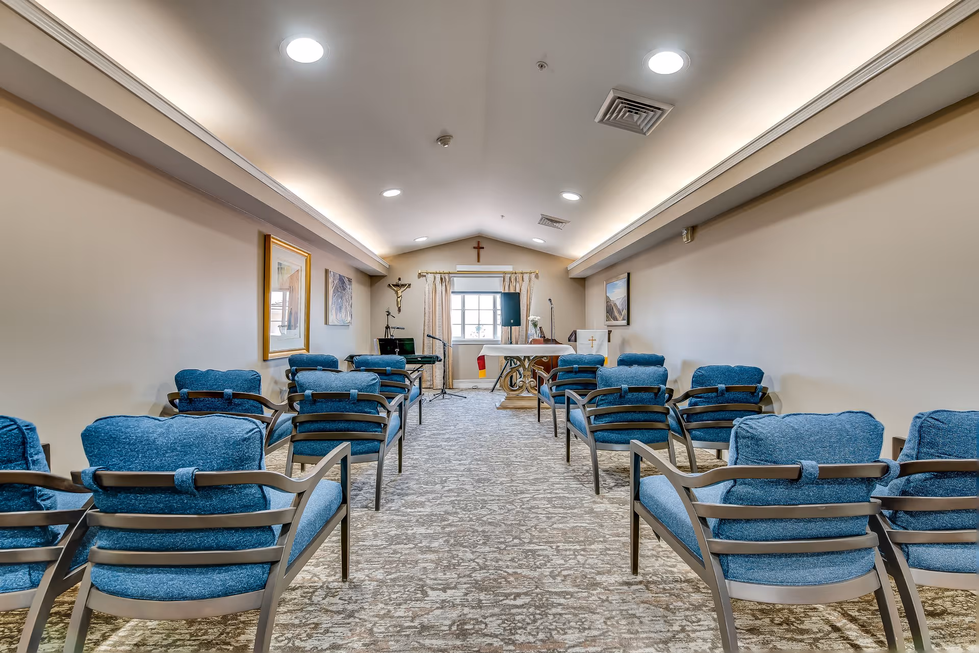 Interior view of a small chapel or worship room with rows of blue cushioned chairs facing an altar at the front. The room has beige walls, framed artwork, a window with curtains, a crucifix on the wall, and a microphone stand near the altar.