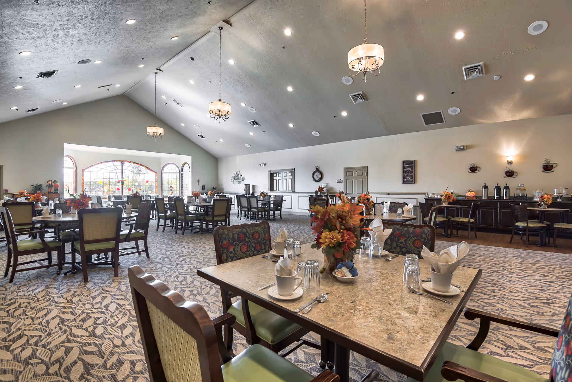 Spacious dining room in an assisted living facility with multiple set tables, floral centerpieces, chandeliers, and large windows.