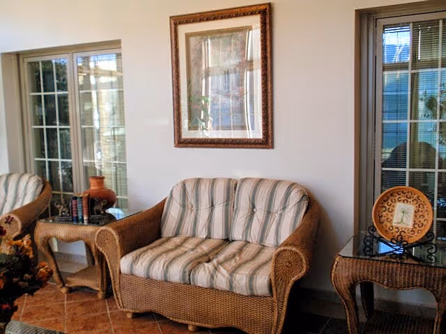 Wicker loveseat with striped cushions flanked by wicker side tables and a framed picture on the wall in a sunlit seating area.