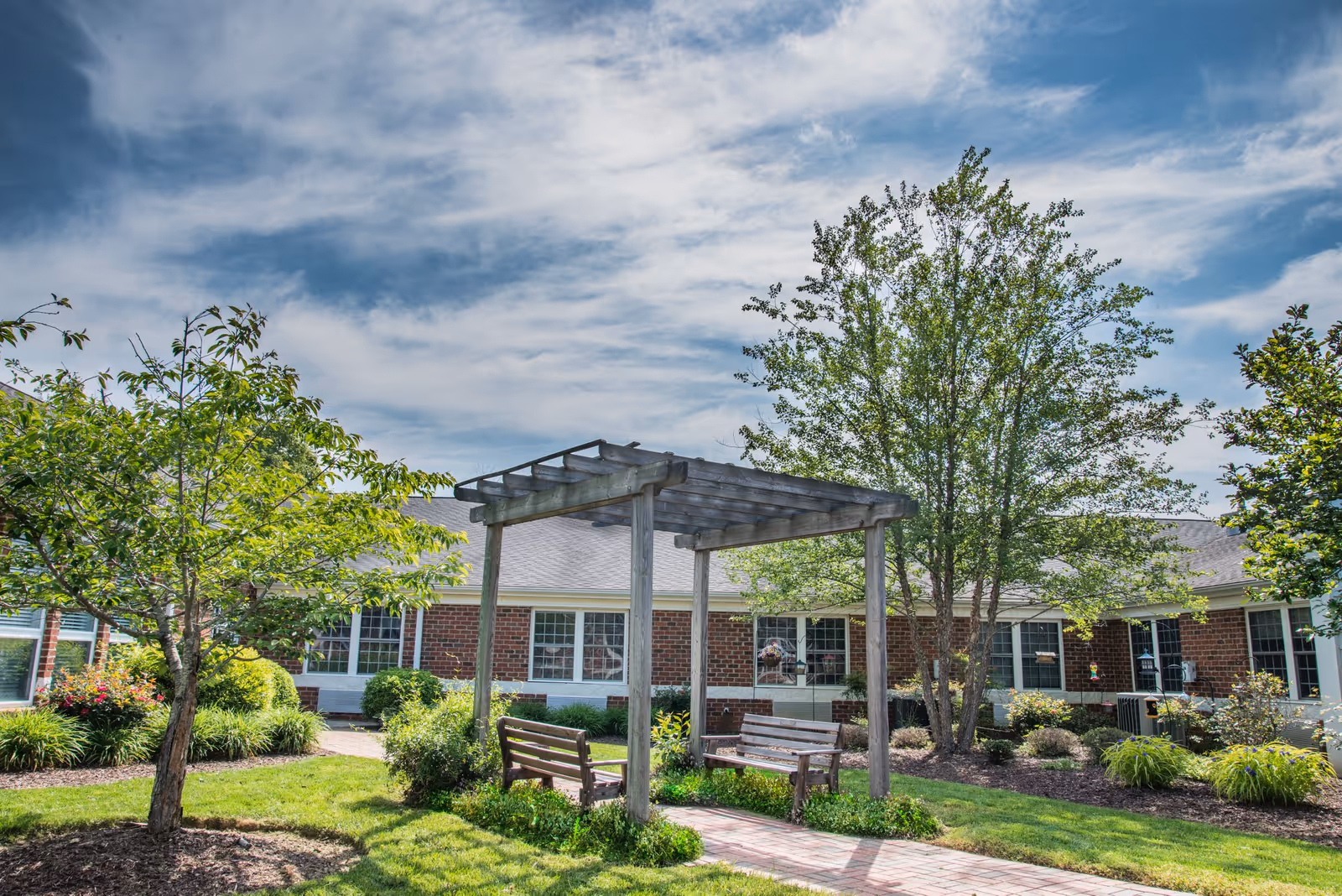 Sunny landscaped courtyard with a wooden pergola, benches, trees and a brick building in the background.