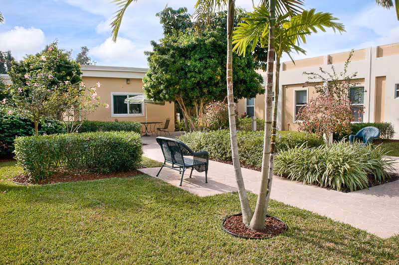 Outdoor garden area at Lakeside Health Center with green grass, palm trees, bushes, and a paved walkway. There are benches and chairs placed along the walkway near the building with beige walls and windows.
