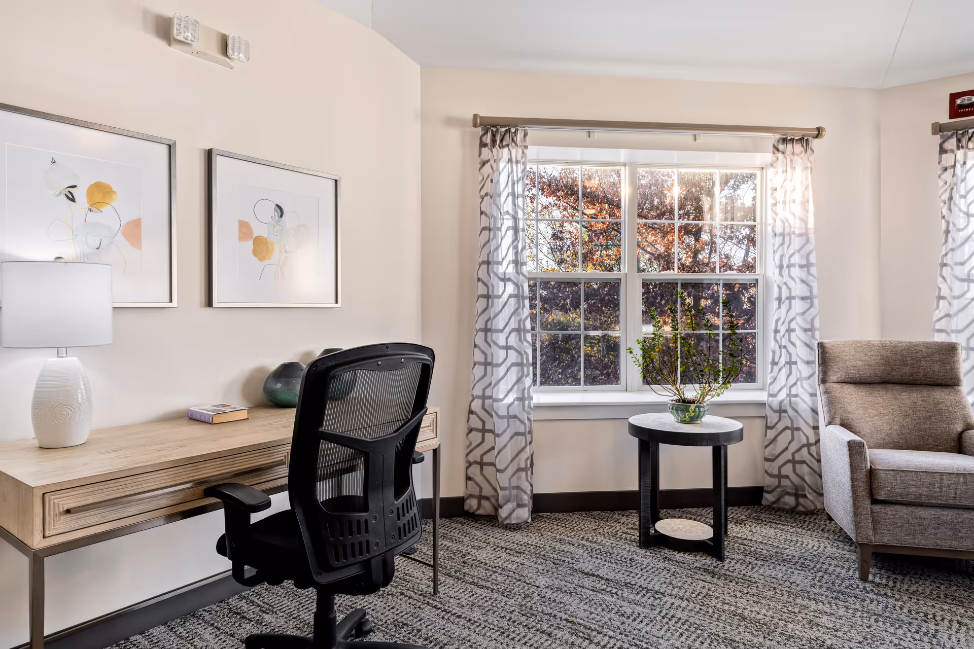 A bright and cozy interior room featuring a wooden desk with a white lamp, a book, and a decorative vase. A black mesh office chair is positioned in front of the desk. Next to a large window with patterned curtains, there is a small round table with a green plant. To the right, there is a comfortable gray armchair. The room has light-colored walls and a patterned carpet.