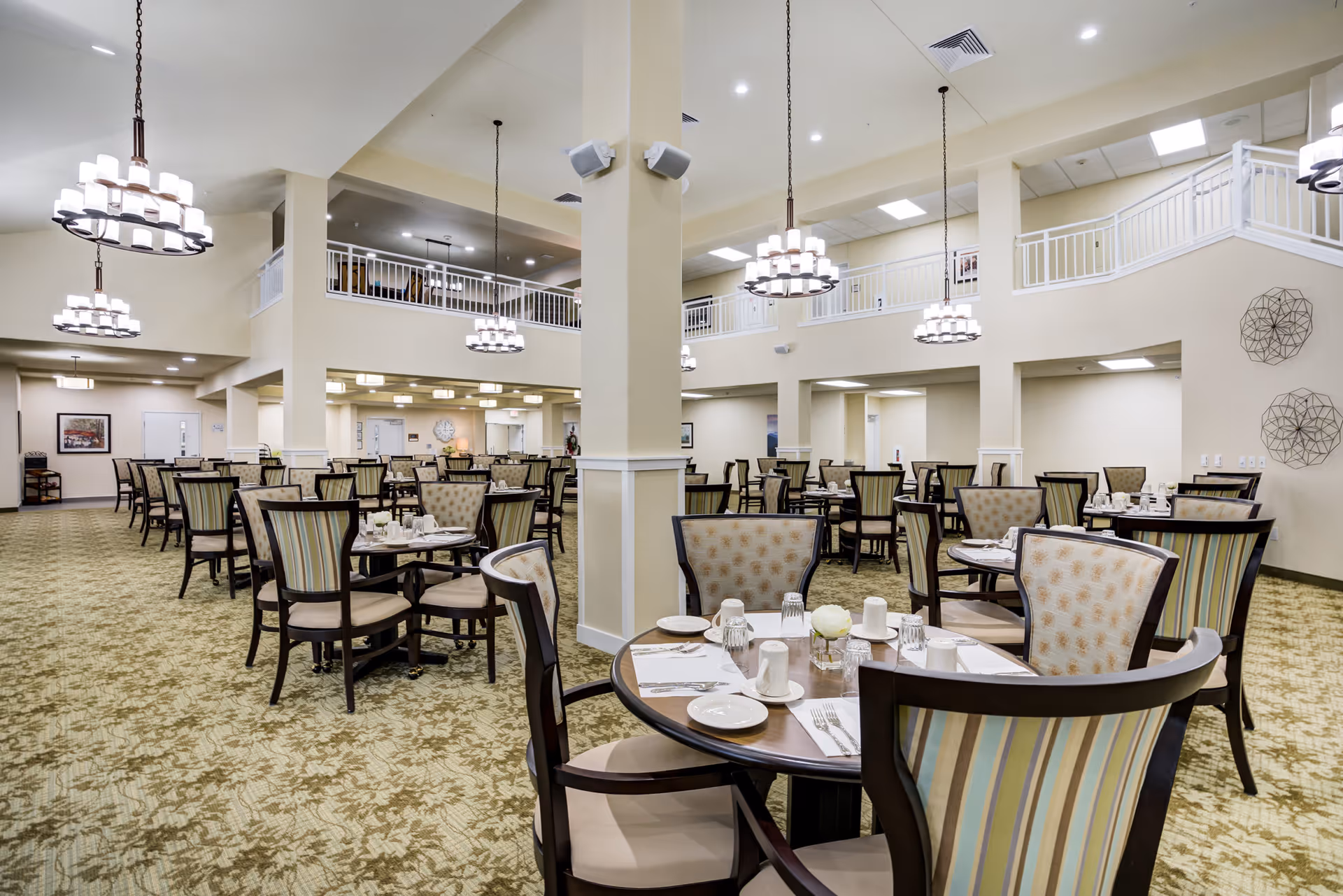 Spacious dining room in a retirement living facility with multiple round tables set with plates, cups, and utensils. The room features high ceilings with hanging chandeliers, patterned carpet, and a second-floor balcony railing overlooking the area.