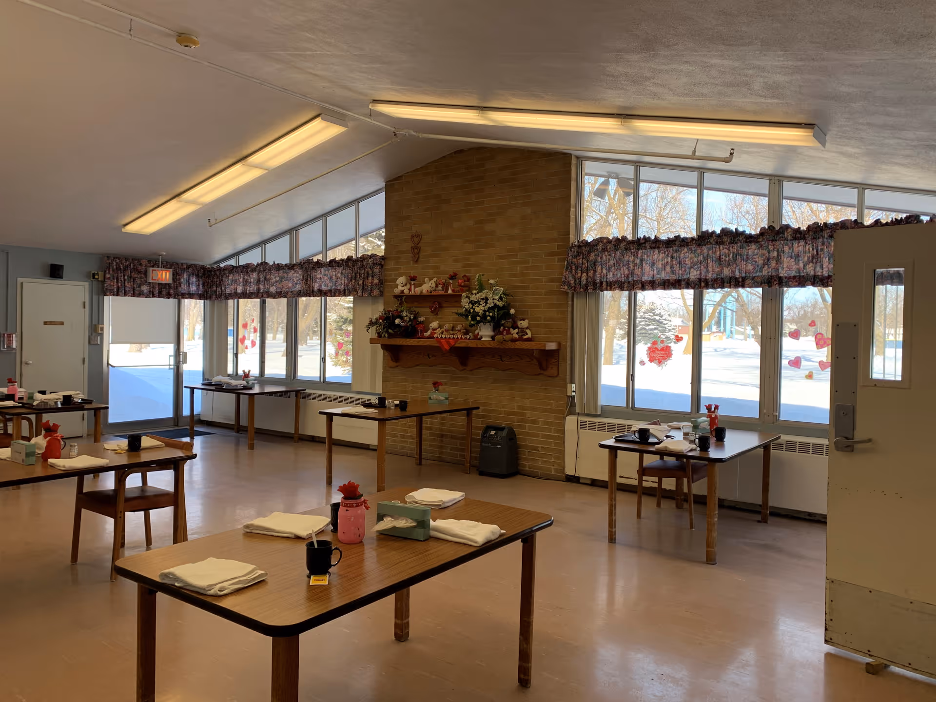 A dining room in a senior living facility with several wooden tables and chairs spaced apart. Each table has a cup, napkins, and some small items. Large windows with floral valances let in natural light and show a snowy outdoor scene. A brick wall with a wooden shelf decorated with flowers and stuffed animals is visible in the background.