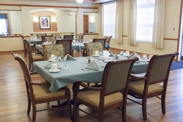 A dining room in a senior living facility with several tables covered with green tablecloths, each set with cups, glasses, napkins, and silverware. The room has wooden floors, beige walls, large windows with curtains, and framed artwork on the walls.