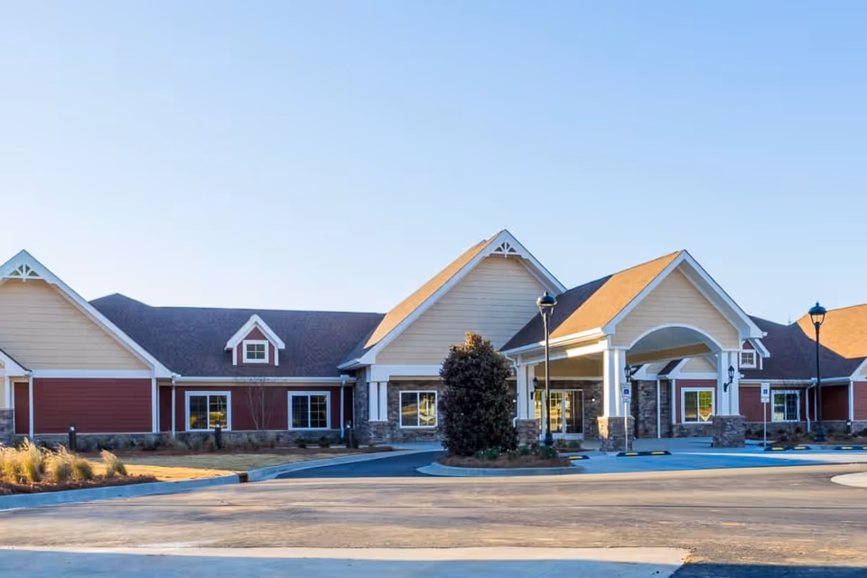 Exterior view of a single-story senior living facility building with a covered entrance, beige and red siding, stone accents, and a clear blue sky.
