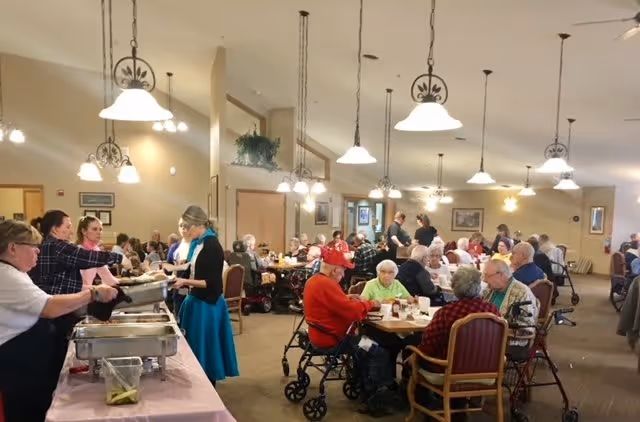 A large dining room filled with elderly people sitting at tables and eating. Several staff members are serving food from buffet trays on the left side of the image. The room is well-lit with hanging pendant lights and ceiling fans, and has a warm, welcoming atmosphere.