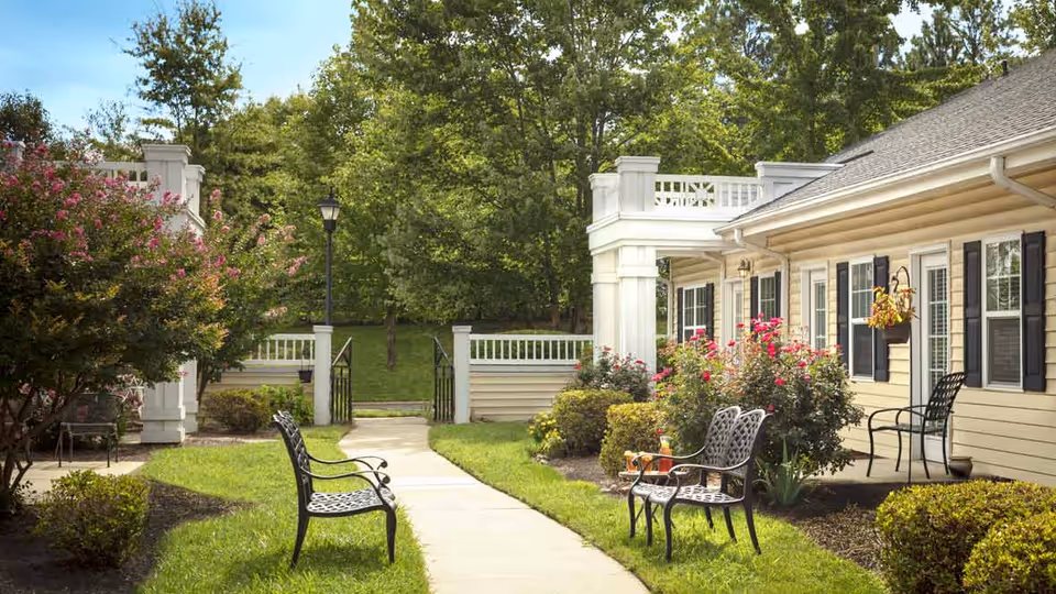 Outdoor garden area at Terrabella Lake Norman featuring a paved walkway, black metal benches, flowering bushes, and a beige building with black shutters and hanging flower pots. Trees and greenery surround the area under a clear blue sky.