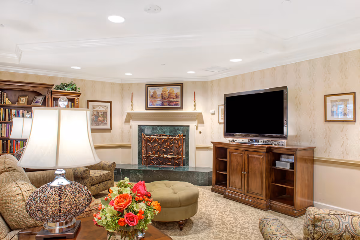 Comfortable living room with sofas, a table lamp and floral centerpiece, an ottoman, fireplace, and a TV on a wooden cabinet.