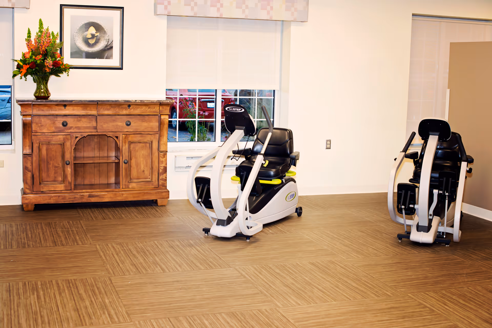 A room with two exercise machines designed for seated workouts, a wooden cabinet with drawers and shelves, a vase with flowers on top, a framed picture on the wall, and windows with blinds partially drawn.