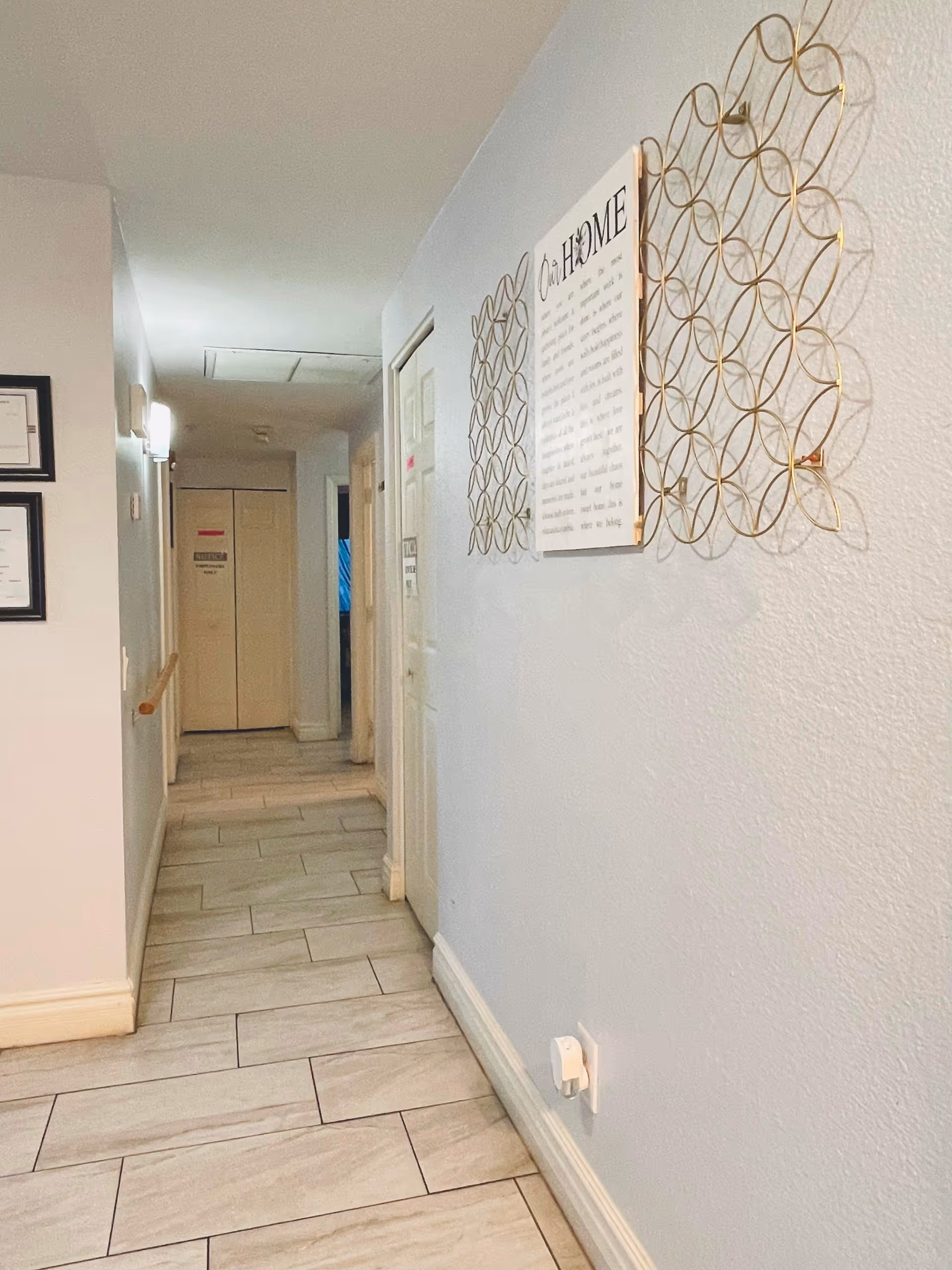 A clean, well-lit hallway with light-colored tiled flooring and light blue walls. On the right wall, there is a decorative gold metal wall art piece with a framed sign titled 'Our Home'. On the left wall, there are framed certificates or documents. The hallway leads to closed doors at the end.