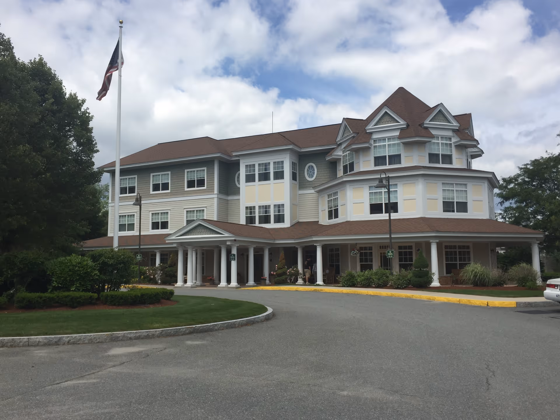 Exterior view of a three-story senior living facility building with a covered entrance supported by white columns, surrounded by landscaped greenery and an American flag on a flagpole in front.
