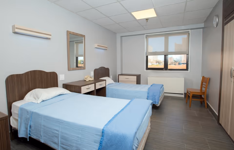 A simple, clean bedroom with two single beds covered in light blue blankets. Between the beds is a wooden nightstand with a vintage telephone and a mirror mounted on the wall above it. There is a window with a roller shade letting in natural light, a wooden chair near the window, and a wardrobe on the right side of the room. The floor is tiled in a dark color and the walls are painted light gray.