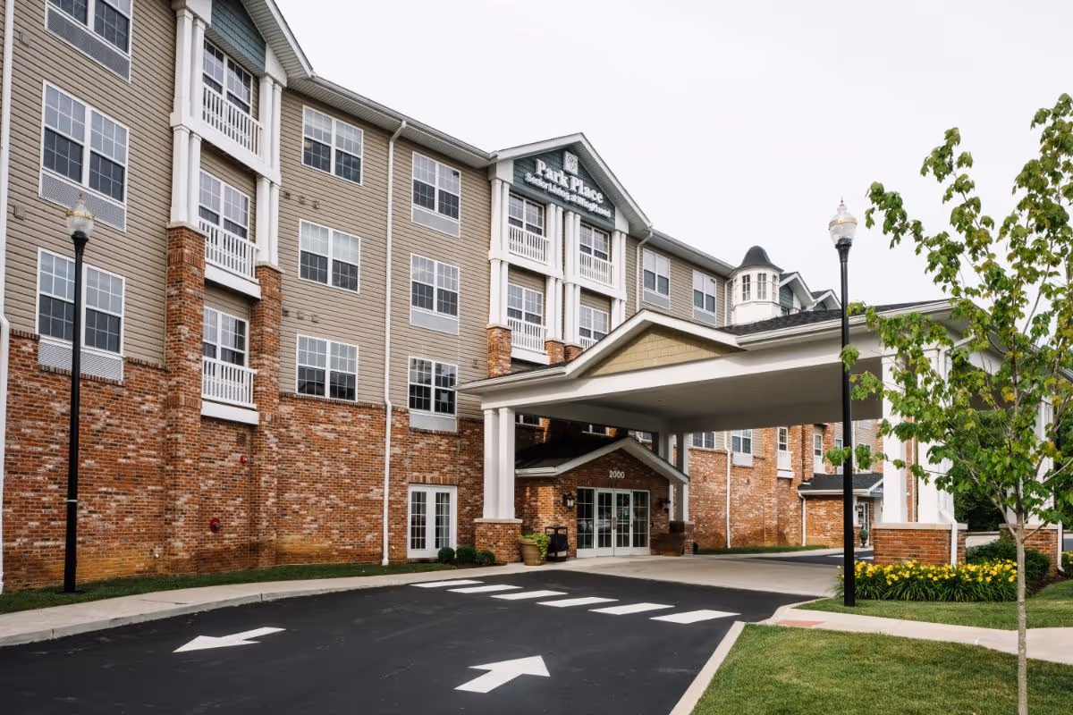 Exterior view of Park Place II senior living facility showing a multi-story building with brick and beige siding, multiple windows, a covered entrance with white pillars, a driveway with directional arrows, street lamps, and landscaped greenery including a tree and yellow flowers.