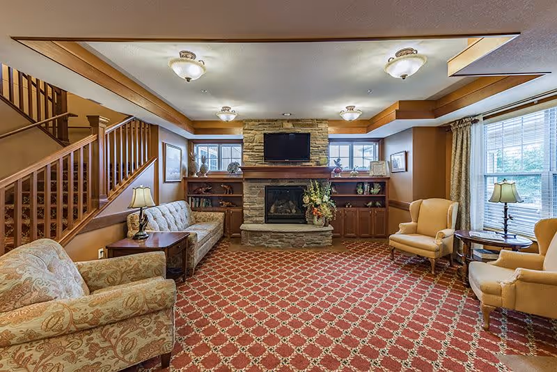 A cozy senior living facility common area with patterned carpet, a stone fireplace with a mounted TV above it, built-in wooden shelves and cabinets, two armchairs, a sofa, side tables with lamps, and large windows with curtains letting in natural light. A staircase with wooden railings is visible on the left side.