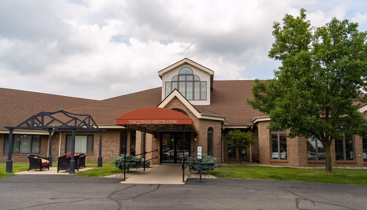 Front exterior view of Tanglewood Assisted Living building with a brick facade, large windows, a red awning over the entrance, a small covered seating area with chairs on the left, and a tree on the right under a cloudy sky.