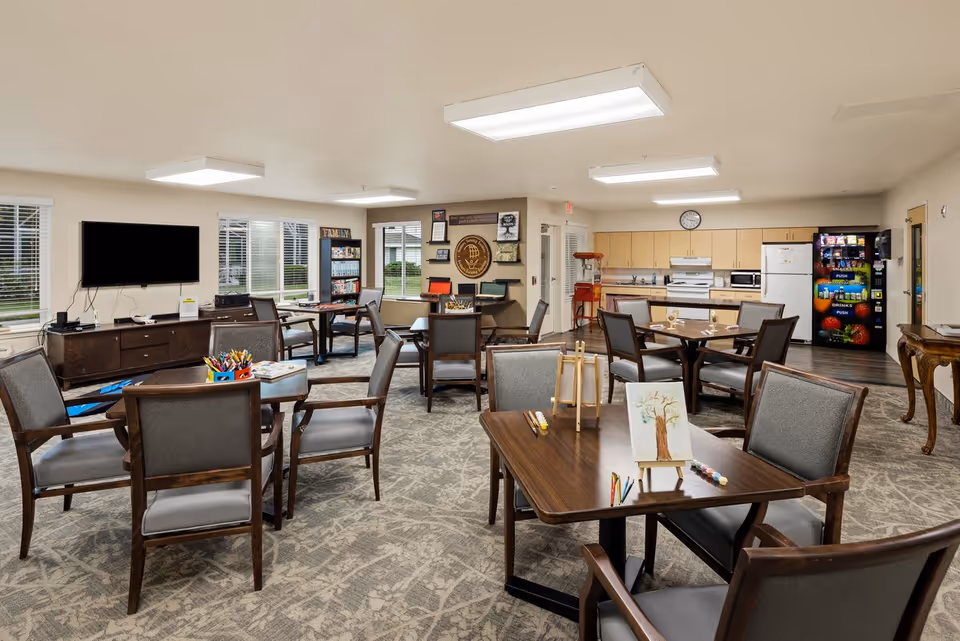 A spacious common area in a senior living community featuring multiple tables and chairs arranged for group activities. One table has art supplies and a small canvas with a tree painting. The room includes a kitchenette with appliances, a vending machine, a popcorn machine, a TV mounted on the wall, and large windows letting in natural light.