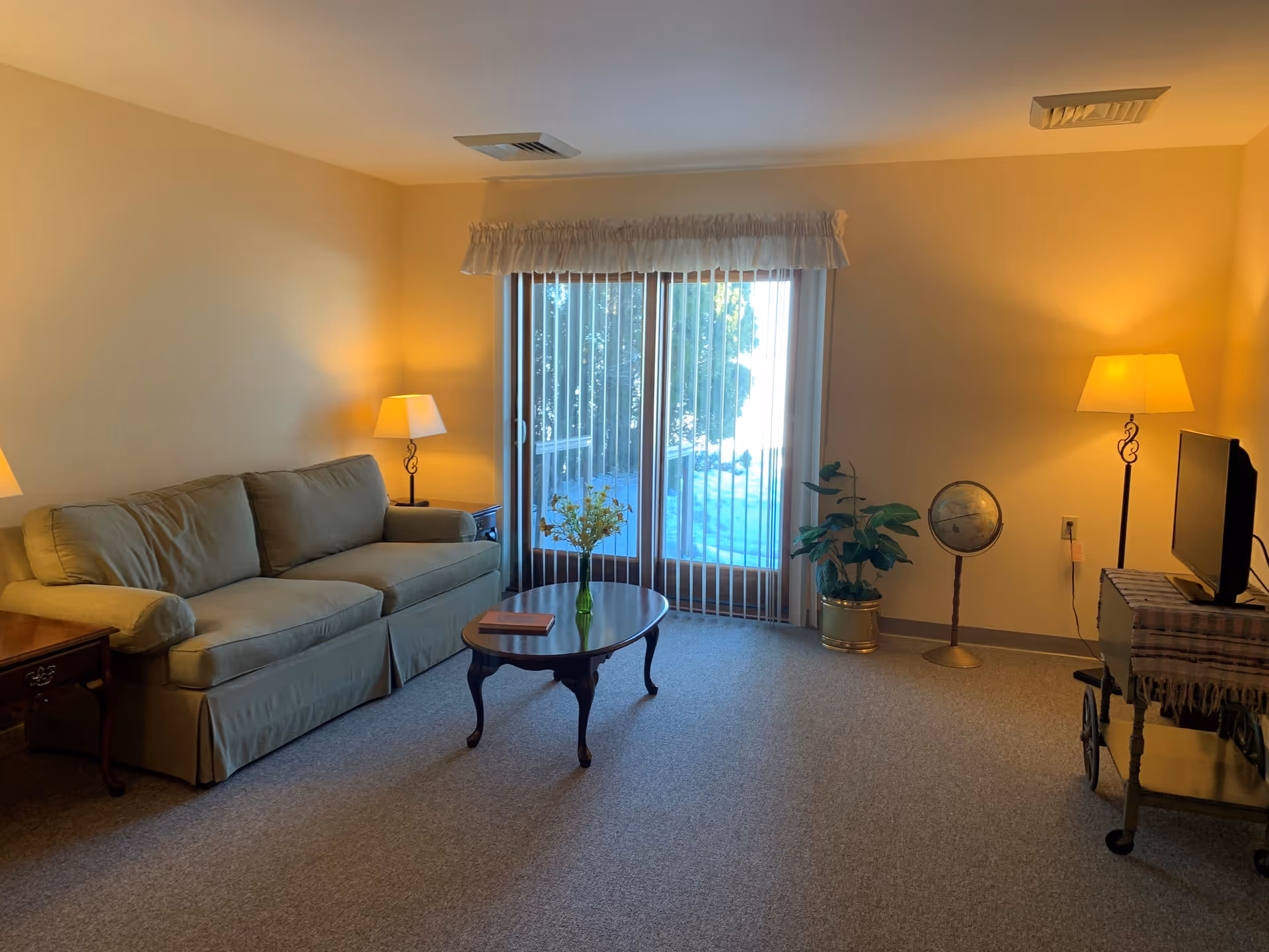 Cozy living room with a beige sofa, wooden coffee table, floor lamps, sliding glass door with vertical blinds, a TV on a cart, and a potted plant.