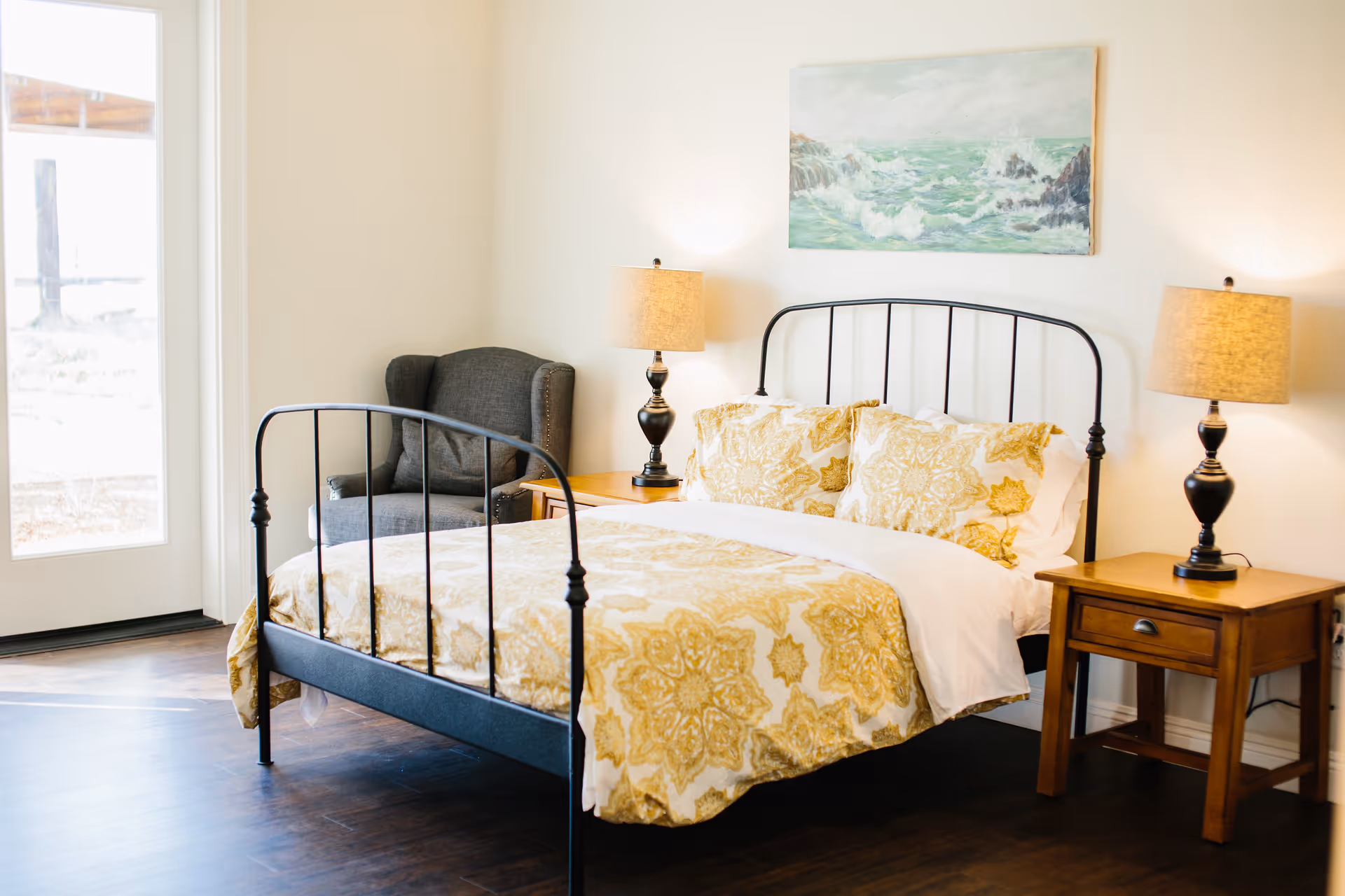 A bright bedroom with a black metal bed frame, yellow patterned bedding, two wooden nightstands each with a lamp, a gray armchair in the corner, and a painting of ocean waves on the wall above the bed. There is a glass door letting in natural light.