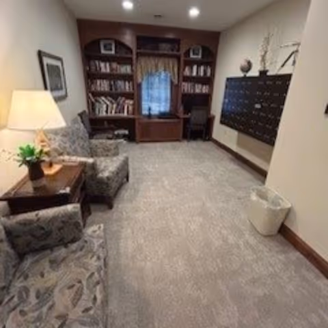 Small cozy sitting room with upholstered chairs, a side table and lamp, bookshelves at the back and mailboxes mounted on the right wall.